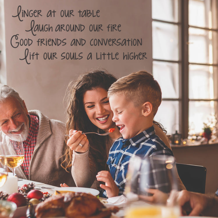 Family enjoying a Thanksgiving or holiday meal with a custom vinyl wall decal above the table that reads 'Linger at our table laugh around our fire Good friends and conversation lift our souls a little higher'. Perfect dining room vinyl lettering for creating a warm, inviting atmosphere, ideal for family gathering wall stickers and holiday decor wall decals.