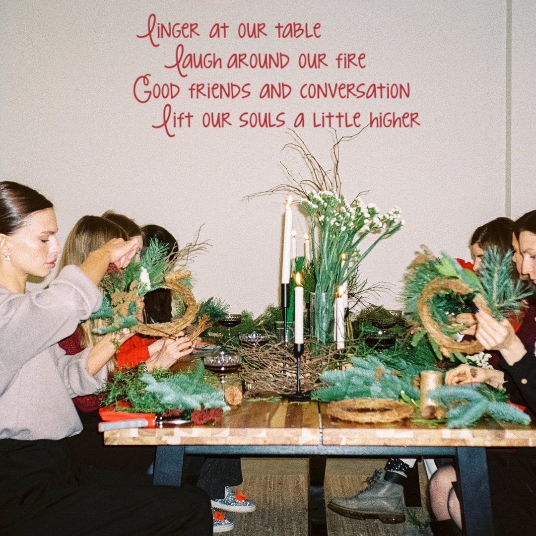 Friends making holiday wreaths at a dining table, with a red vinyl wall quote 'Linger at our table laugh around our fire Good friends and conversation lift our souls a little higher' on the wall. This removable wall decor is perfect for DIY craft space inspiration or Christmas party wall stickers.