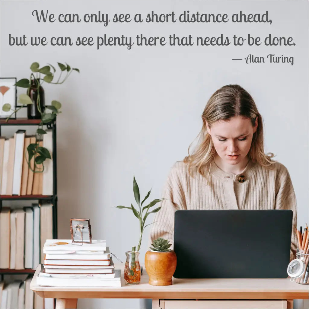 A woman sitting at a desk with a laptop, surrounded by books and a plant, with a customizable vinyl wall decal of Alan Turing's quote displayed on the wall.