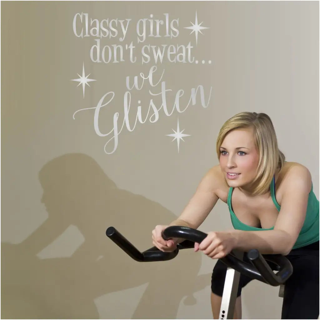 Pretty girl on exercise bike in front of a large wall decal in the gym that reads: Classy girls don't sweat; we glisten.