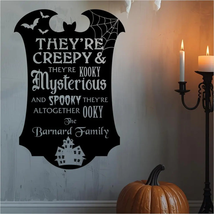 A black "Creepy & Kooky" personalized Halloween wall decal with bats and a haunted house, displayed on a lightly textured wall next to a pumpkin and lit candelabra.