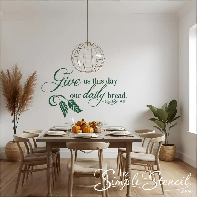 A warm dining room featuring a large green "Give us this day our daily bread. Matthew 6:11" wall decal above a wooden table set for a meal. The elegant script decal with a wheat design is highlighted by a modern spherical chandelier, creating a blessed and grateful atmosphere for Thanksgiving or daily family meals.
