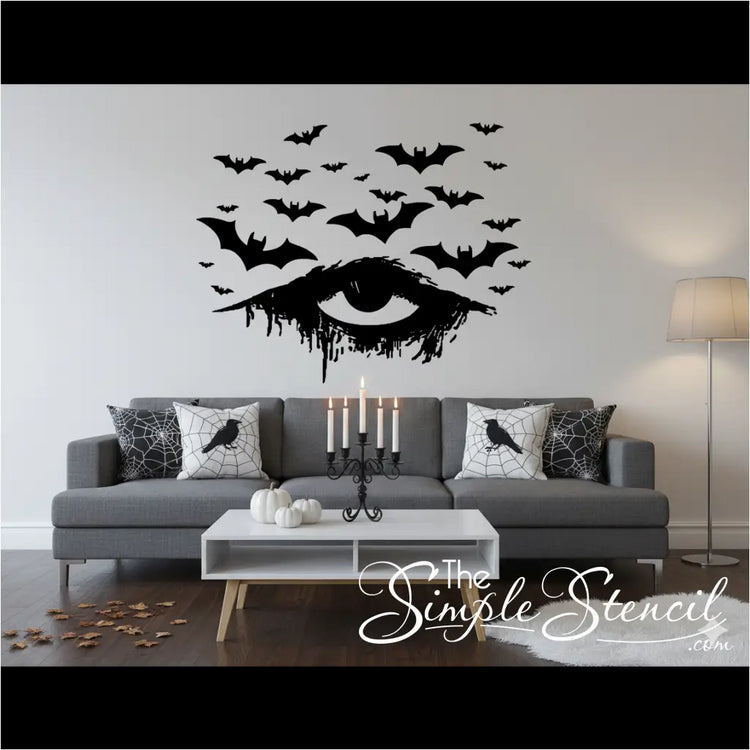 A black vinyl Halloween decal of a distressed, dripping eye with numerous bats flying above it, applied to a clean white wall in a stylish living room. The room features a gray sofa, white pumpkins, a black candelabra, and pillows with crow and spiderweb designs, creating a minimalist yet sophisticated Halloween aesthetic.