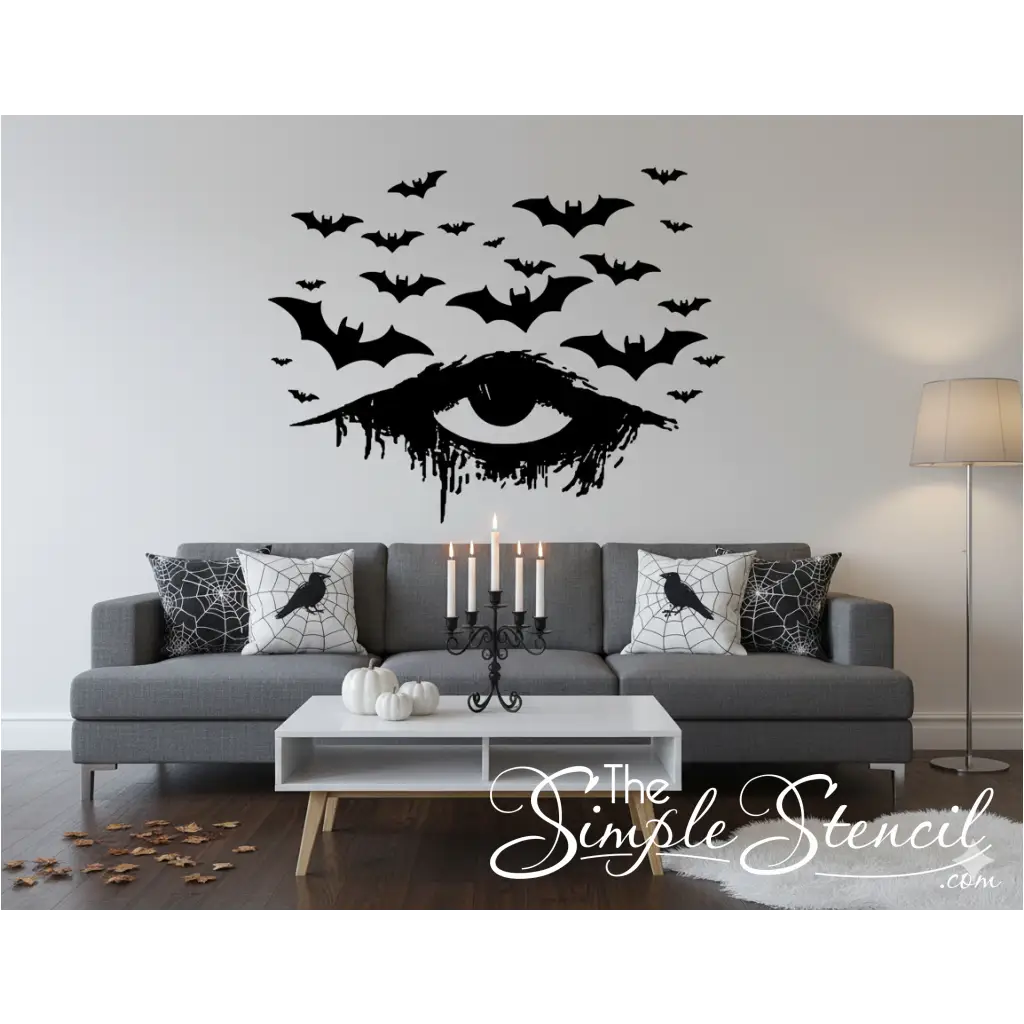 A black vinyl Halloween decal of a distressed, dripping eye with numerous bats flying above it, applied to a clean white wall in a stylish living room. The room features a gray sofa, white pumpkins, a black candelabra, and pillows with crow and spiderweb designs, creating a minimalist yet sophisticated Halloween aesthetic.