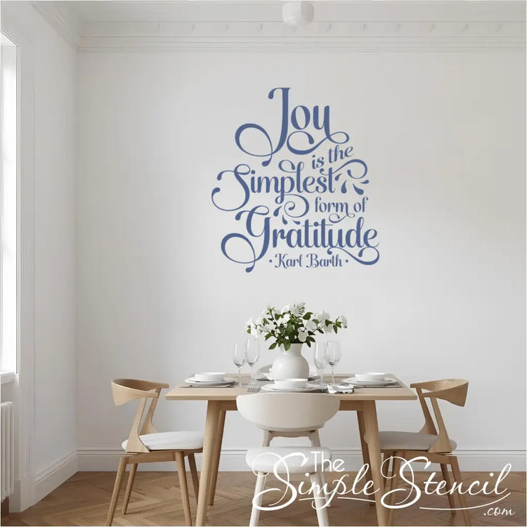 Elegant dining room featuring a large "Joy is the Simplest Form of Gratitude" wall decal in a beautiful blue script font. The decal is centered above a modern wooden dining table set with white dishes, glasses, and a vase of white flowers, creating a serene and grateful atmosphere perfect for Thanksgiving home decor or everyday dining room inspiration.
