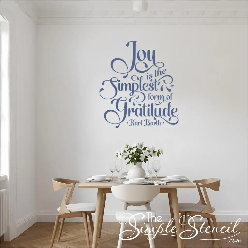 Elegant dining room featuring a large "Joy is the Simplest Form of Gratitude" wall decal in a beautiful blue script font. The decal is centered above a modern wooden dining table set with white dishes, glasses, and a vase of white flowers, creating a serene and grateful atmosphere perfect for Thanksgiving home decor or everyday dining room inspiration.
