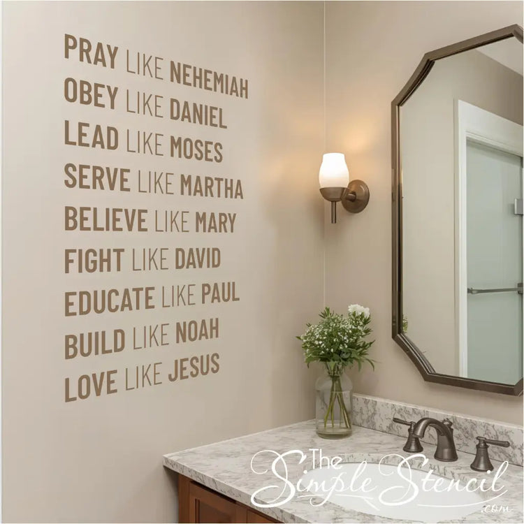 A brown wall decal listing nine biblical virtues, including "PRAY LIKE NEHEMIAH" and "LOVE LIKE JESUS," installed on a light beige wall above a bathroom sink. The scene includes a marble countertop, a bronze-framed mirror, and a vase with flowers.