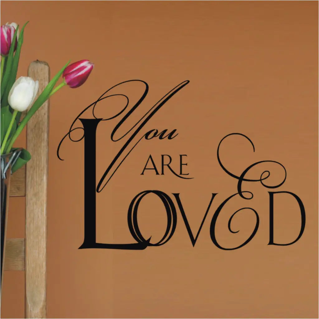 Romantic script design of a vinyl wall decal that reads YOU ARE LOVED and looks great installed on a wall near a flower arrangement where recipient will feel loved every single time they look at this beautiful wall art.