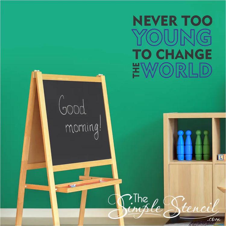 A colorful "Never Too Young to Change the World" vinyl decal displayed on a bright classroom wall with children's artwork. 