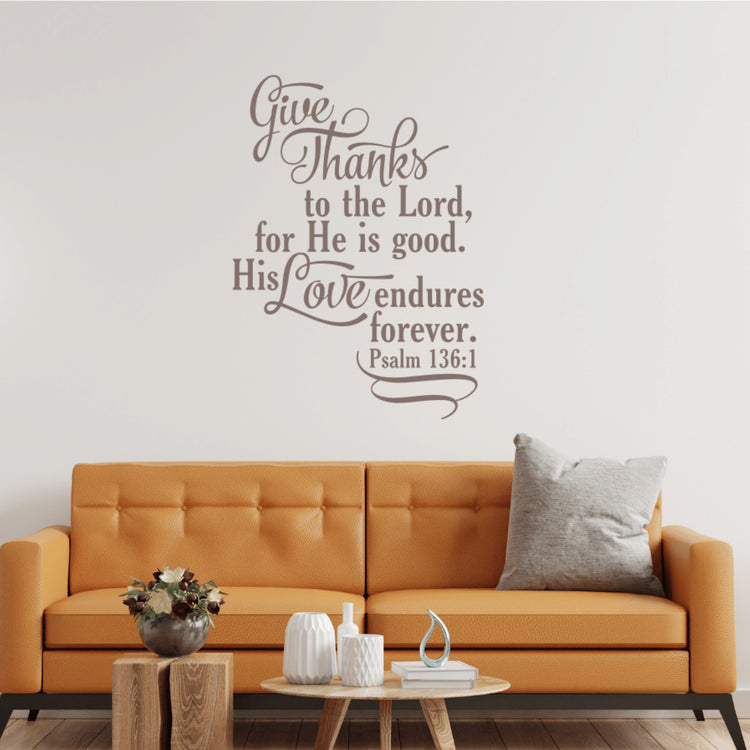 Psalm 136:1 "Give Thanks to the Lord" vinyl decal in taupe, installed above a modern orange living room couch with throw pillows.