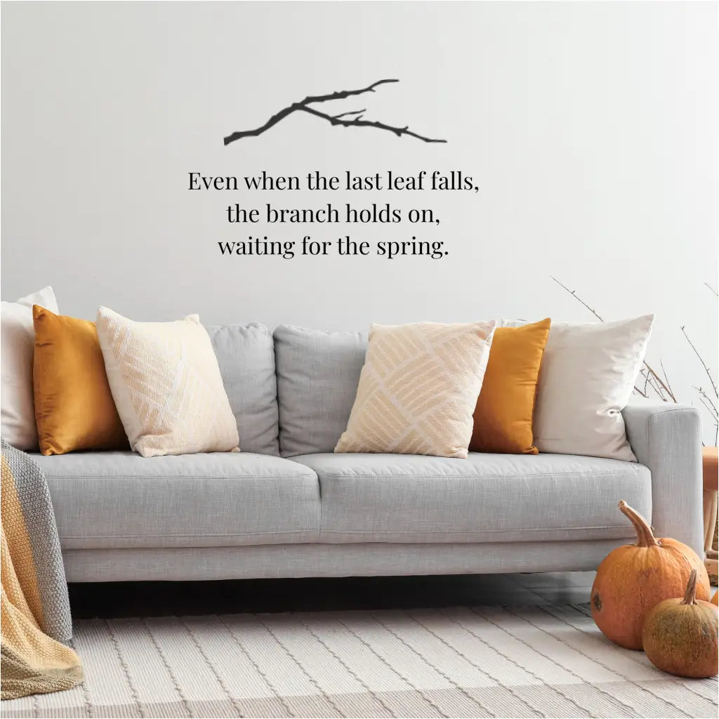 A black bare branch vinyl wall decal paired with the quote "Even when the last leaf falls, the branch holds on, waiting for the spring" above a cozy grey sofa with autumn-colored pillows.