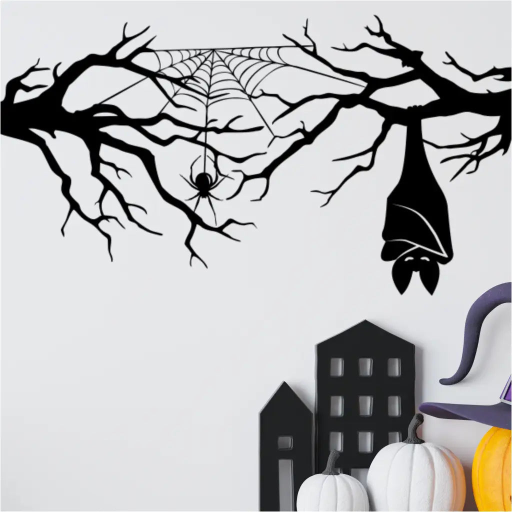 A black Halloween decal of a tree branch, spider web, and hanging bat applied to a white wall, serving as a backdrop for white and orange pumpkins and a purple witch's hat, creating a charming indoor Halloween display.