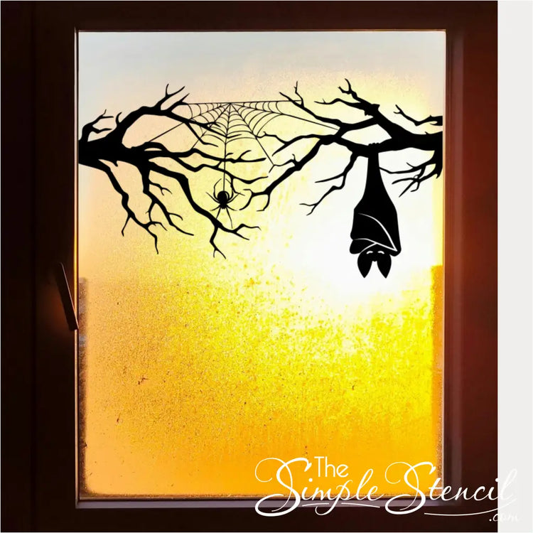 A Halloween window decal featuring a black silhouette of a gnarled branch, spider web, and hanging bat, set against a vibrant orange and yellow sunset view through the window. By TheSimpleStencil
