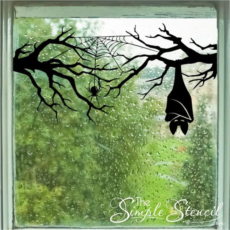 A black vinyl Halloween Simple Stencil decal of a tree branch, spider web, and hanging bat applied to a rainy glass window, creating a spooky and atmospheric scene with green foliage outside.