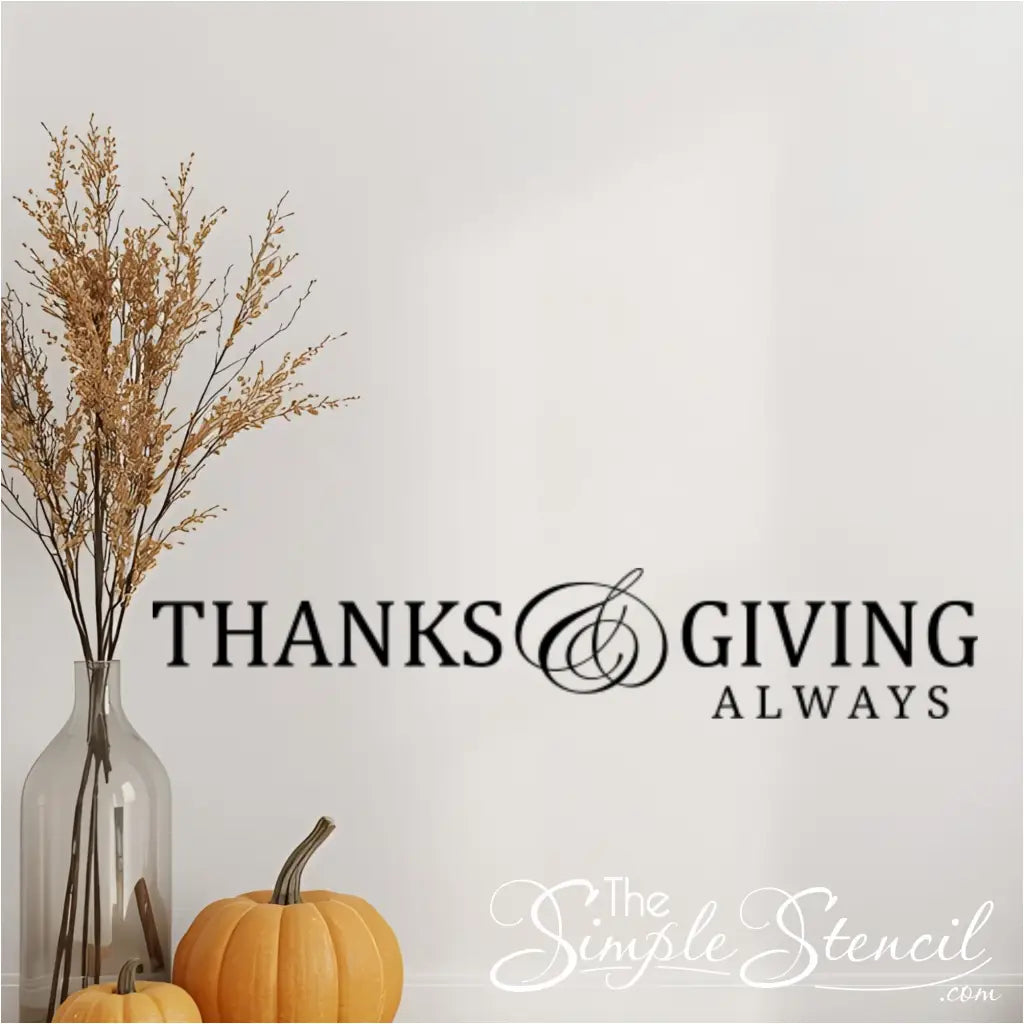 A 'Thanks and Giving Always' vinyl wall sticker shown above a small table with two pumpkins and a vase of dried botanicals, representing seasonal fall and Thanksgiving decor.