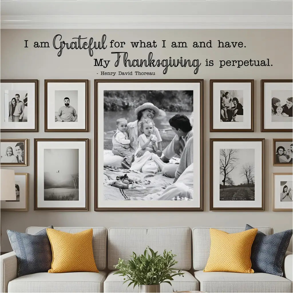 A living room with a cozy sofa and a large gallery wall of black and white family photos. The "I am grateful for what I am and have..." wall decal is placed as a central, guiding piece of art for the entire photo collection, highlighting its use in creating a permanent, gratitude-focused display.