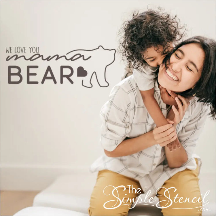 "We Love You Mama Bear" wall decal in a warm brown vinyl applied to a living room wall, with a cozy armchair and throw pillows nearby a mom and son playing. By The Simple Stencil