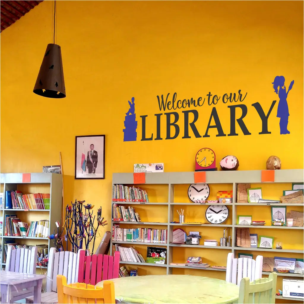 A school library with a bright yellow wall featuring a decal sign with the text 'Welcome to our LIBRARY' and silhouettes of a boy and girl reading.