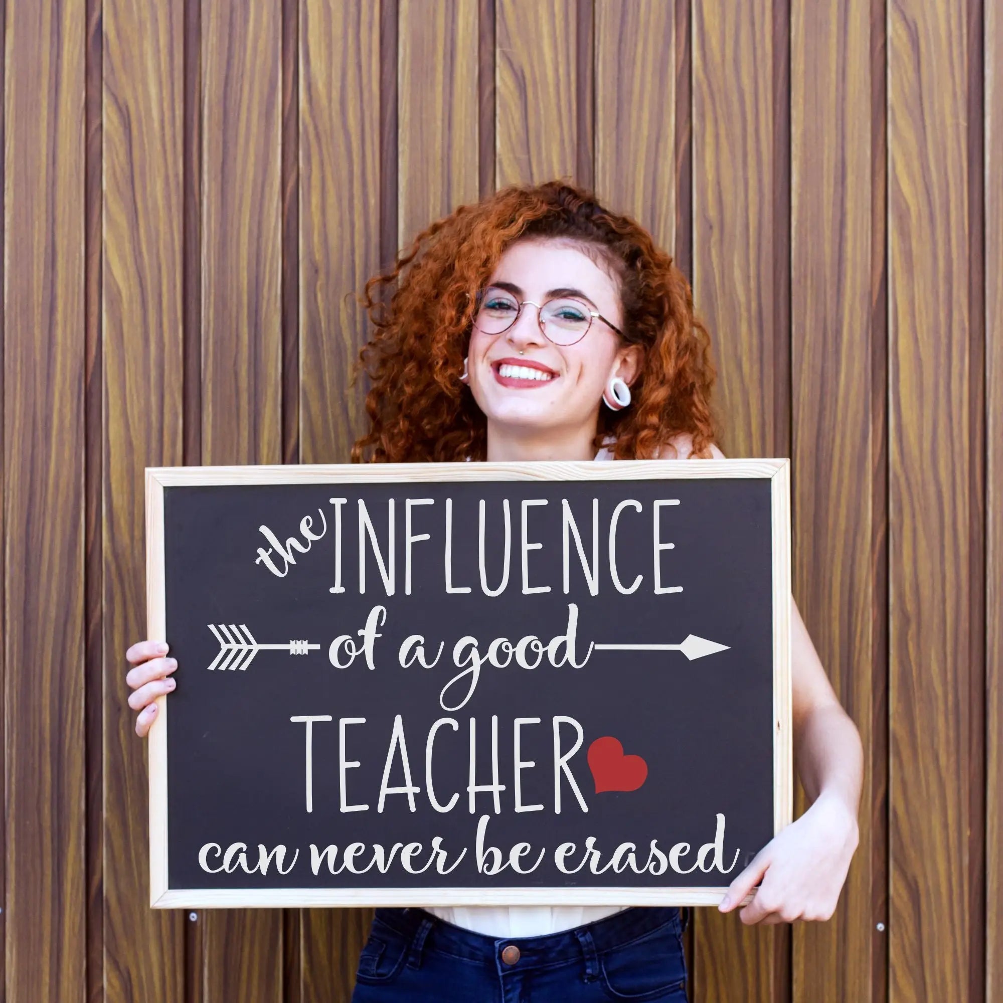 A happy teachers displaying a sign her students gifted her during teacher appreciation week that reads: The influence of a good teacher can never be erased. By TheSimpleStencil