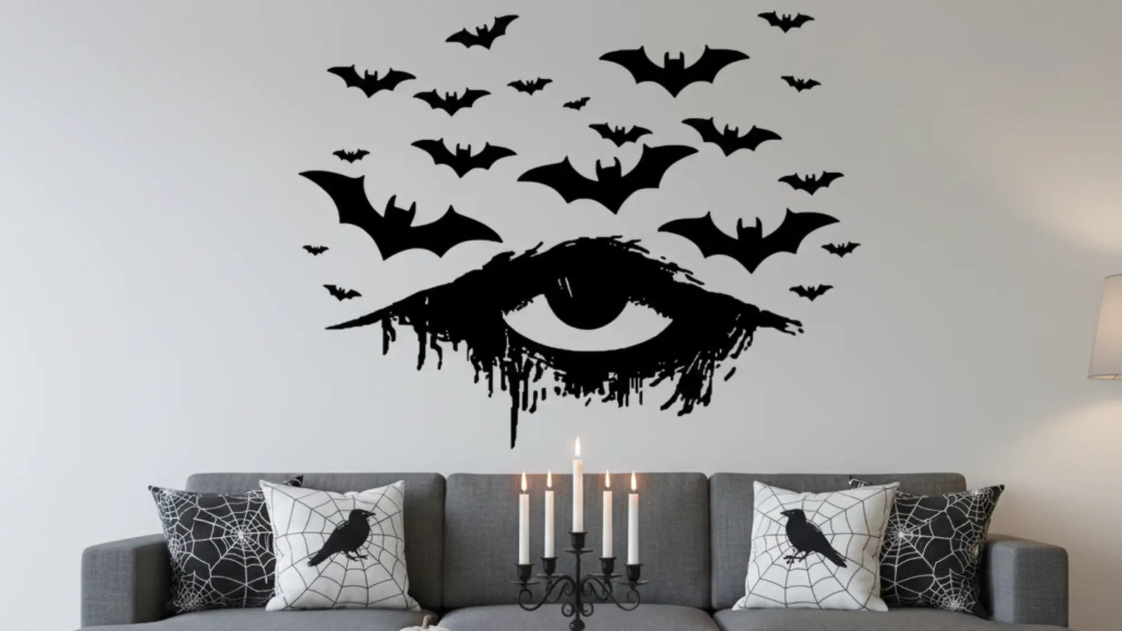 Halloween living room with a large black dripping gazing eye wall decal surrounded by bats above a gray sofa, styled with spiderweb and crow pillows. This sophisticated, eerie decor inspires high-end, unique Halloween transformations.