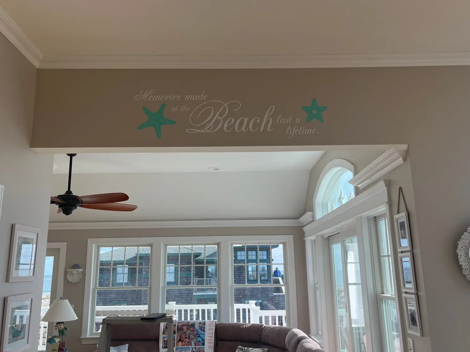 A sun-drenched beach panorama adorns a cozy living room wall, framed by a custom decal reading "Memories made at the beach last a lifetime." Starfish add a touch of coastal charm to this personalized beach house decor.