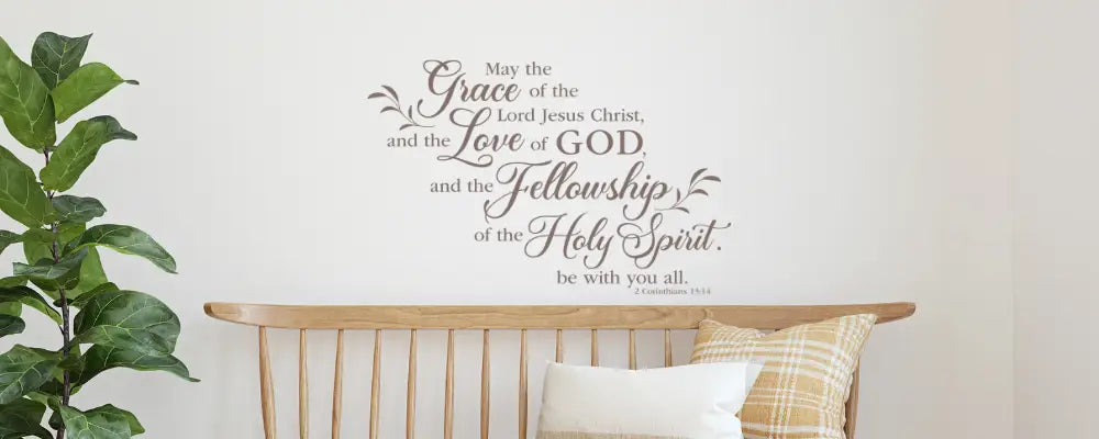 Large-scale Corinthians 13:14 wall decal featuring the 'Grace of the Lord Jesus Christ' in an elegant, flowing script, displayed in a bright living room above a wooden bench and potted plant.