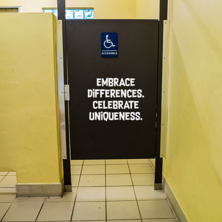 White vinyl decal on a black school restroom stall door reading Embrace Differences Celebrate Uniqueness for a positive bathroom transformation project.