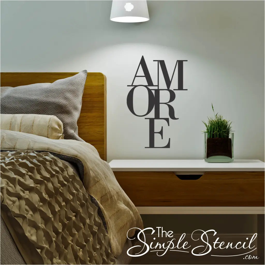 Dark grey stacked 'AMORE' wall decal shown above a modern white and wood nightstand with a small plant, next to a bed featuring textured beige bedding. Master suite accent piece.