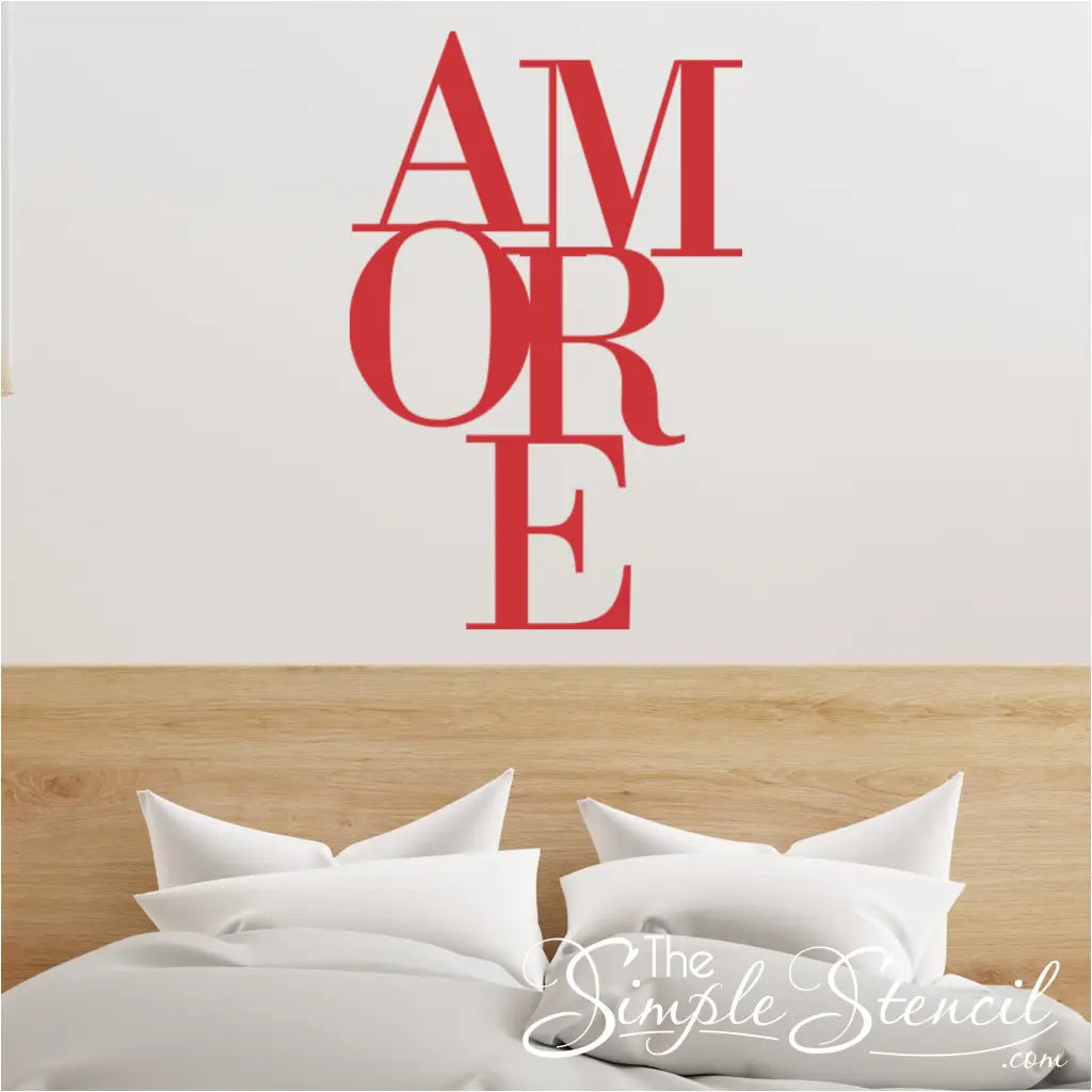 Red typography wall art decal reading 'AMORE' in a stacked serif font, placed above a light oak wooden headboard and white bedding. Ideal Valentine's Day decor.