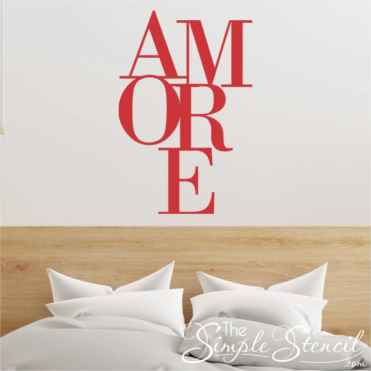 Red typography wall art decal reading 'AMORE' in a stacked serif font, placed above a light oak wooden headboard and white bedding. Ideal Valentine's Day decor.