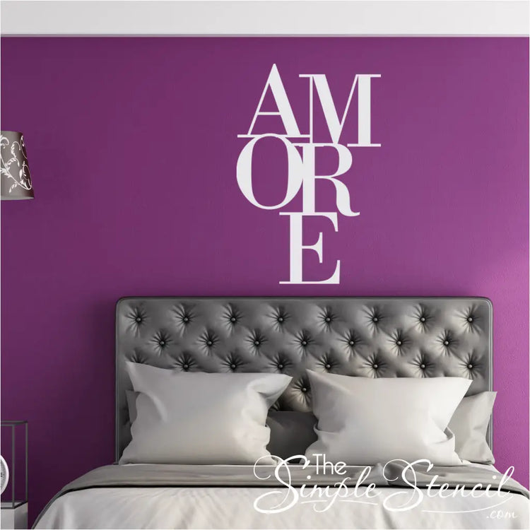 White stacked 'AMORE' vinyl wall decal centered above a grey tufted headboard in a master bedroom with deep purple walls. Romantic bedroom decor idea.