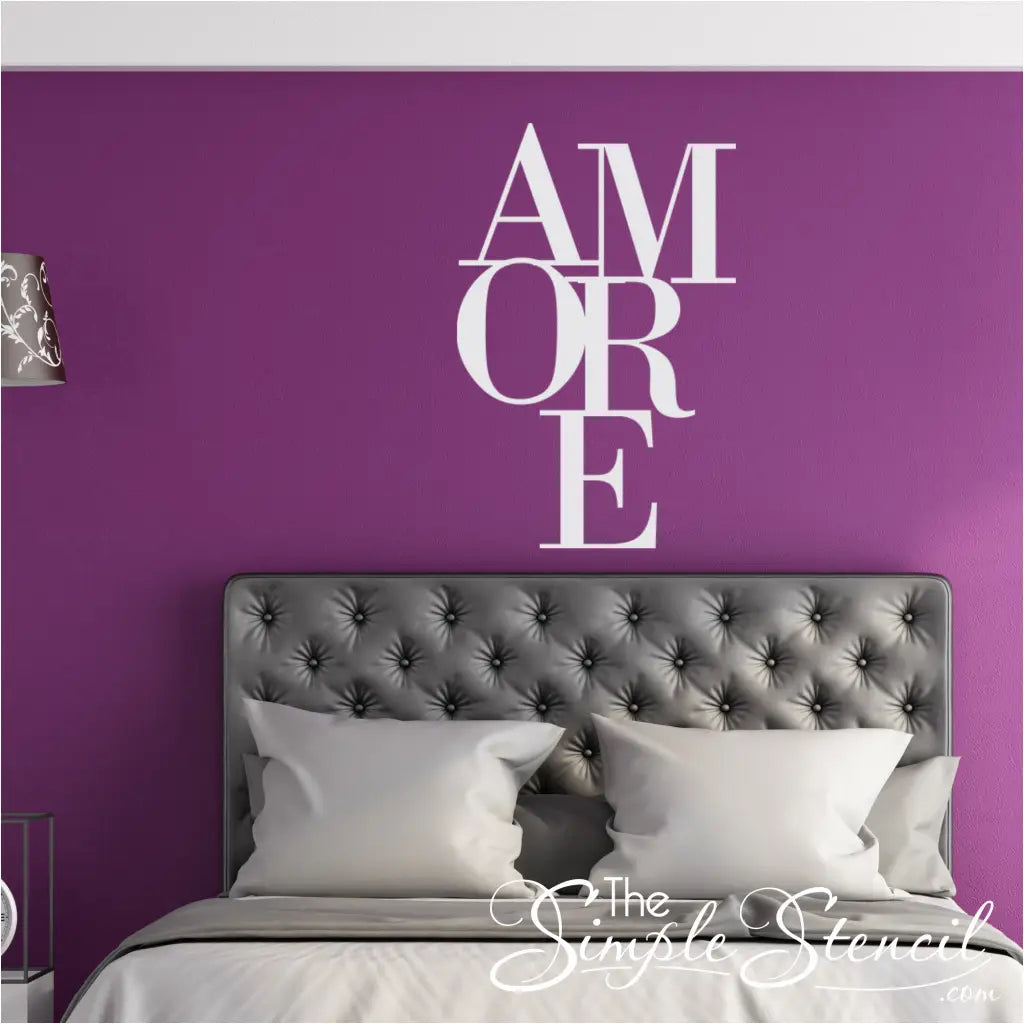 White stacked 'AMORE' vinyl wall decal centered above a grey tufted headboard in a master bedroom with deep purple walls. Romantic bedroom decor idea.