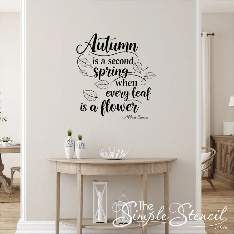 Decorative wall quote 'Autumn is a second spring when every leaf is a flower' on a neutral wall with a wooden table and decor items.