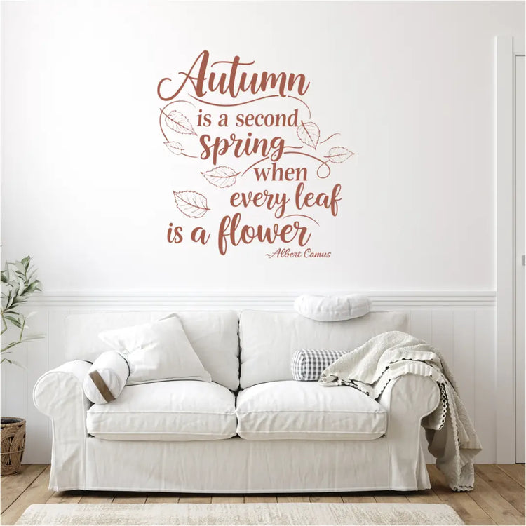 A brown wall decal with the quote "Autumn is a second spring when every leaf is a flower." by Albert Camus, installed above a white sofa in a modern living room with a rug and a plant.