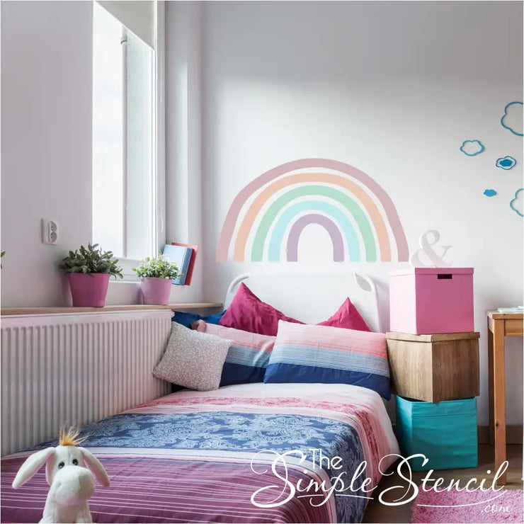 Sweet pastel colored rainbow wall decal shown behind a girls bed frame creates a fun look that instantly lifts the rooms vibes with colorful fun decor by The Simple Stencil