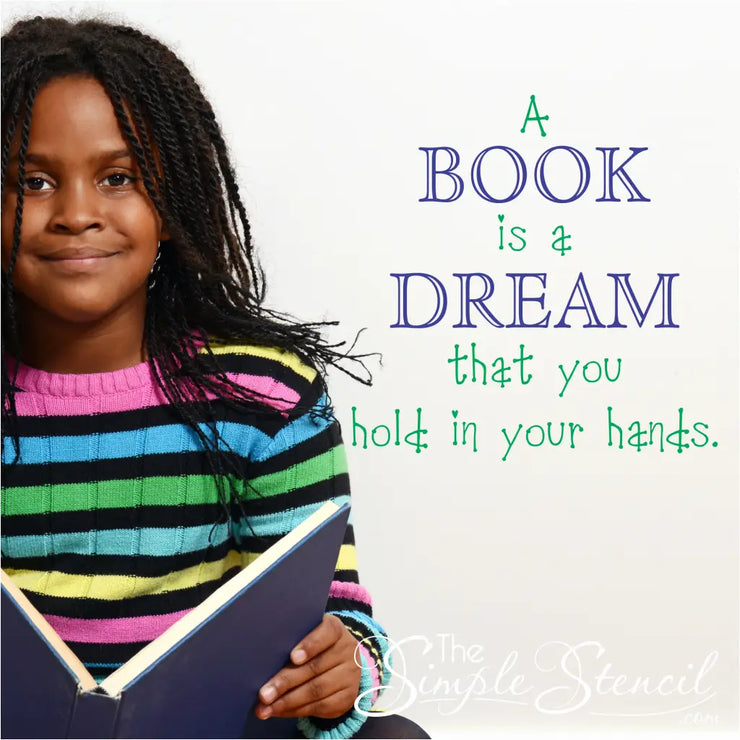 A young elementary school student reading a book next to a library wall decal by TheSimpleStencil.com that reads: A BOOK is a DREAM that you hold in your hands.