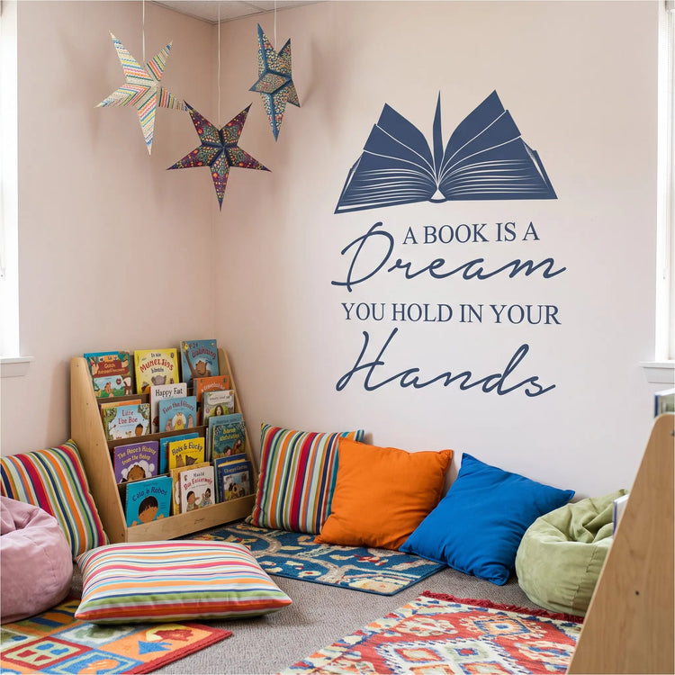 A colorful primary school reading corner featuring the decal customized in a dark blue color. Natural lighting from windows highlights the hand-painted, matte finish on the soft pastel accent wall. Below the smaller-scale decal sits a variety of oversized floor cushions, playful bean bags, and low-level shelving holding childrens' books.