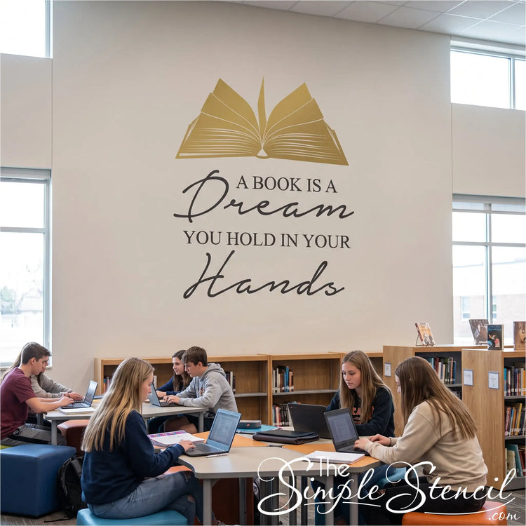 A large black vinyl wall decal with a gold open book illustration and the quote 'A BOOK IS A Dream YOU HOLD IN YOUR Hands' is displayed on a beige library wall. Several students are seated at tables working on laptops in the background of the library.