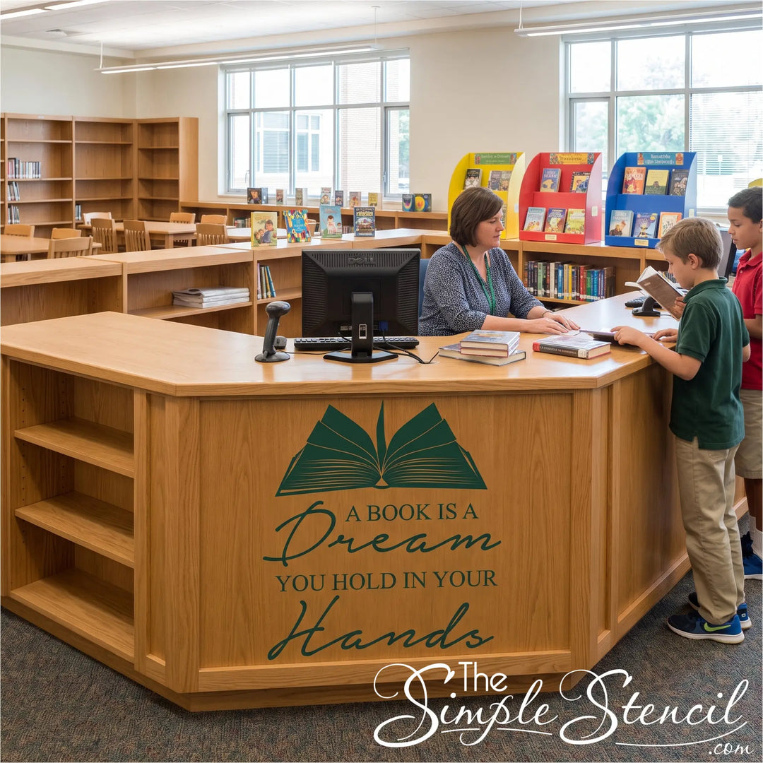 A large wooden library desk with shelves and a computer station, featuring the printed quote 'A BOOK IS A Dream YOU HOLD IN YOUR Hands' with an open book graphic. A librarian sits behind the desk while two children interact with the computer in a bright, modern library.