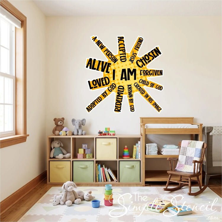 A photograph of a vibrant children's playroom and children's ministry room. On a light blue wall, a large, yellow and black "I AM" sun-burst affirmation wall decal is the central focal point. The text on the decal includes phrases like "I AM," "A NEW PERSON," "CHILD OF GOD," and "LOVED." Below the decal are white toy storage shelves with colorful bins. In the corner, wooden floating shelves display children's books and stuffed animals (a teddy bear and an elephant). On the wood floor are patterned play mats