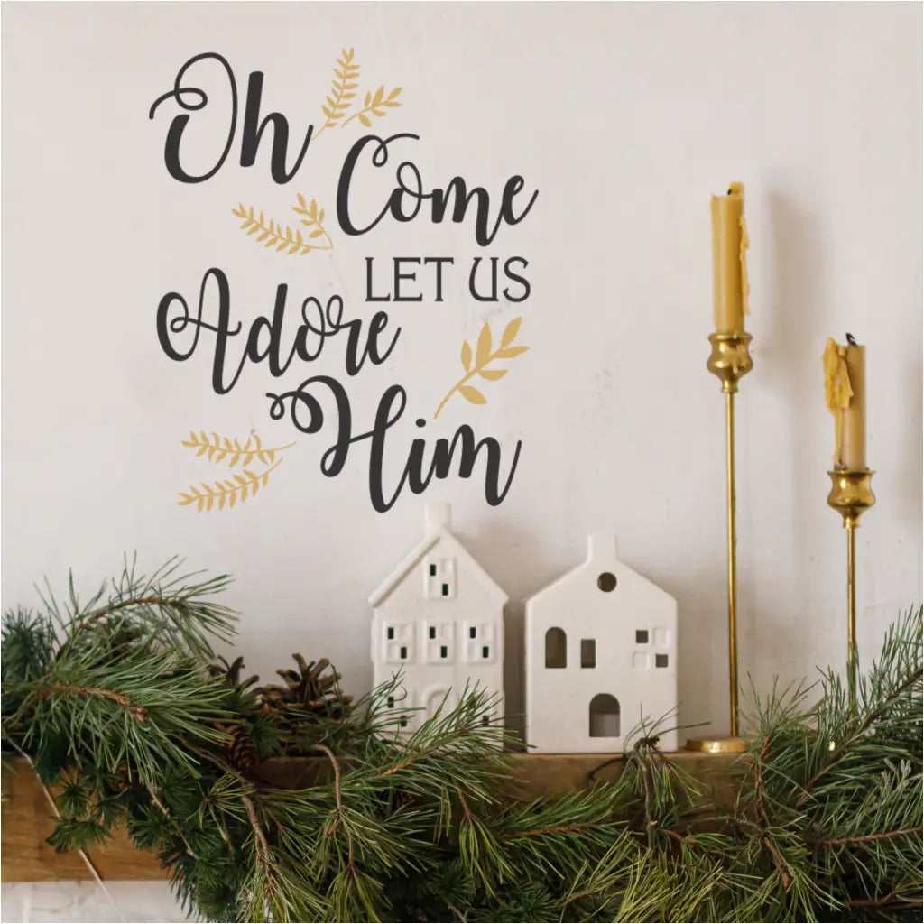 A stylish holiday mantle decorated with pine garland, white ceramic houses, and golden candlesticks, featuring a prominent black and gold "Oh Come Let Us Adore Him" wall decal on the wall above. Ideal removable Christmas wall stickers for festive home decoration.