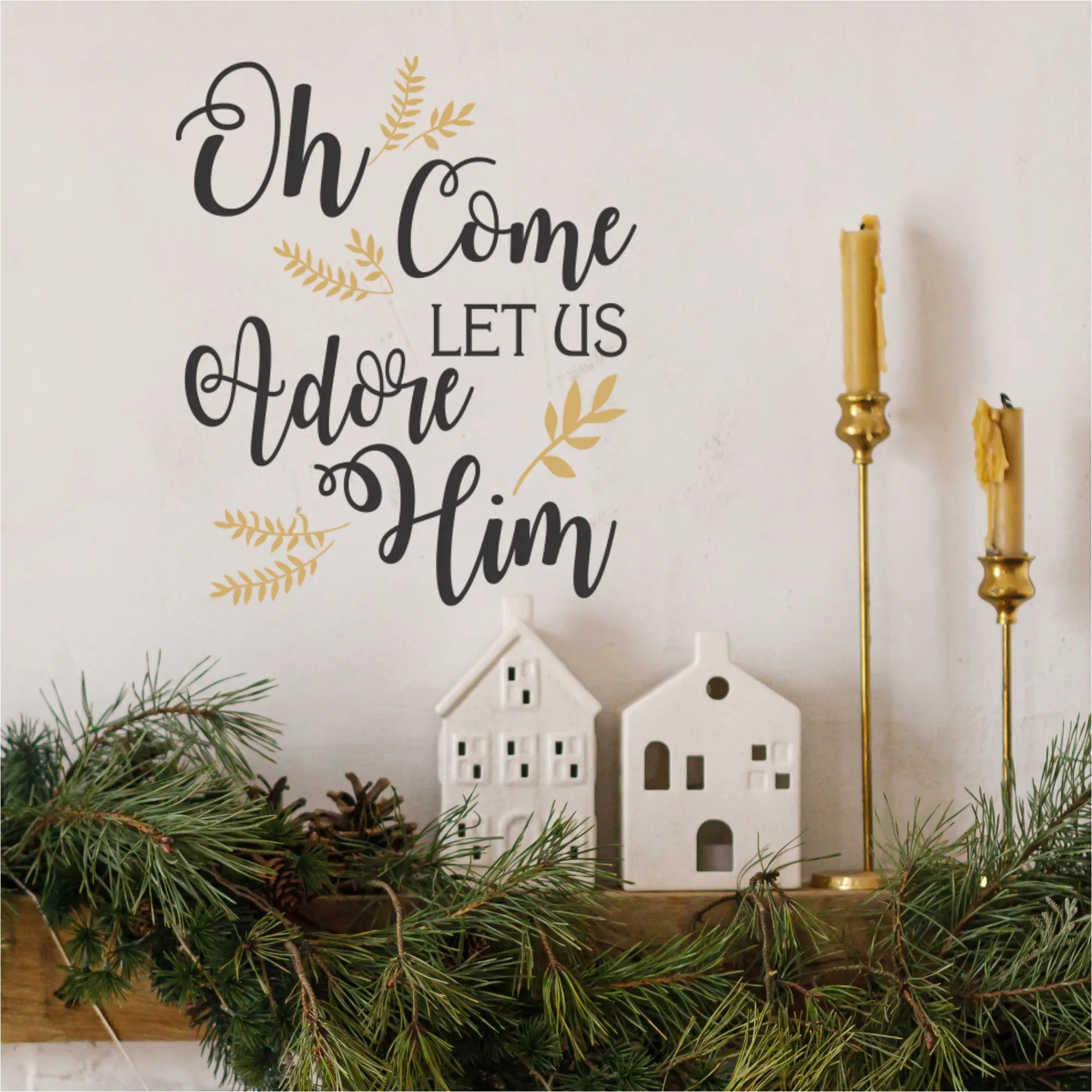 A stylish holiday mantle decorated with pine garland, white ceramic houses, and golden candlesticks, featuring a prominent black and gold "Oh Come Let Us Adore Him" wall decal on the wall above. Ideal removable Christmas wall stickers for festive home decoration.