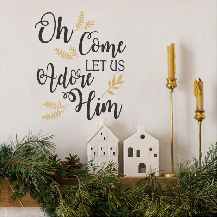 A stylish holiday mantle decorated with pine garland, white ceramic houses, and golden candlesticks, featuring a prominent black and gold "Oh Come Let Us Adore Him" wall decal on the wall above. Ideal removable Christmas wall stickers for festive home decoration.