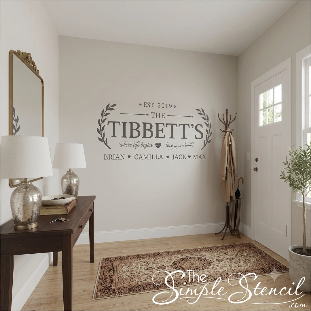 A styled entryway with a personalized wall decal reading 'THE TIBETT'S' and names below, mounted on a light neutral wall. The scene includes a wooden console table with two white table lamps, a gold-framed mirror, a coat rack with a beige coat, a patterned area rug, light wood flooring, and a potted plant near a white door.