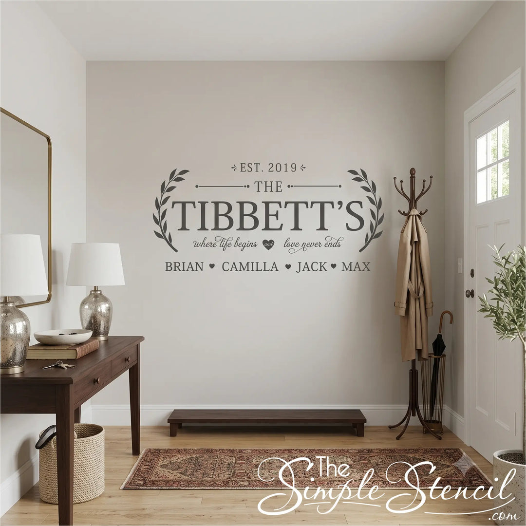 A square photo of a bright, modern home entryway featuring a custom charcoal grey family name monogram decal centered on a warm beige wall. The decal reads 'EST. 2019 THE TIBBETT'S' with a family quote and names at the bottom. A dark wood console table with gold lamps sits to the left, a vintage-style Persian rug is on the wood floor, and a classic coat rack stands near the front door.
