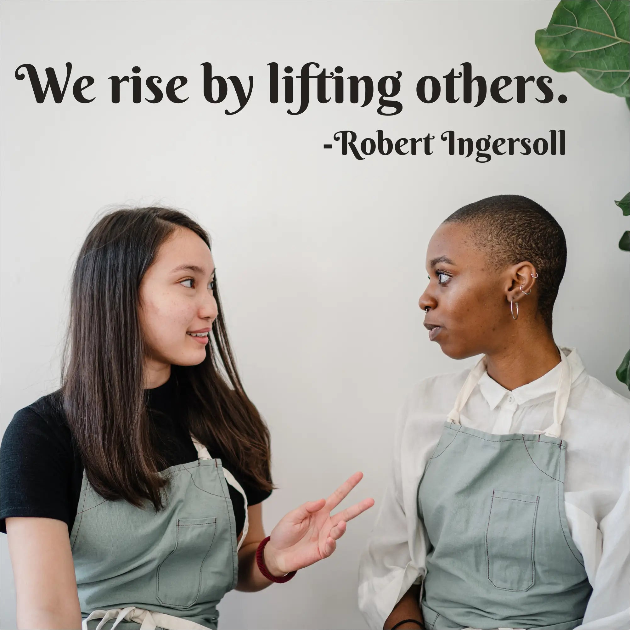 Two women wearing matching light blue aprons pose beside a customizable we rise by lifting others vinyl wall decal
