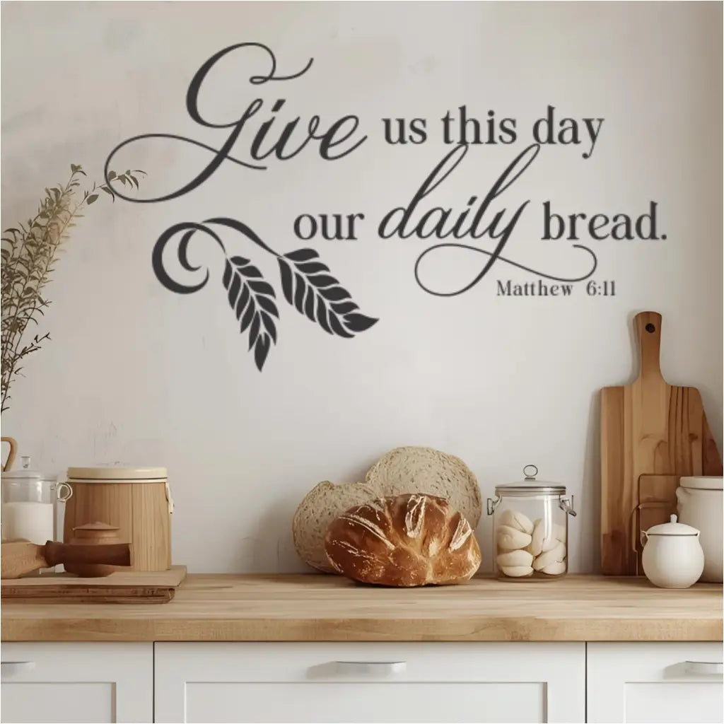 A rustic farmhouse kitchen scene featuring a black "Give us this day our daily bread. Matthew 6:11" wall decal above a wooden countertop. The decal is surrounded by fresh bread, kitchen canisters, and wooden cutting boards, emphasizing a blessing over food and a grateful spirit in the heart of the home.