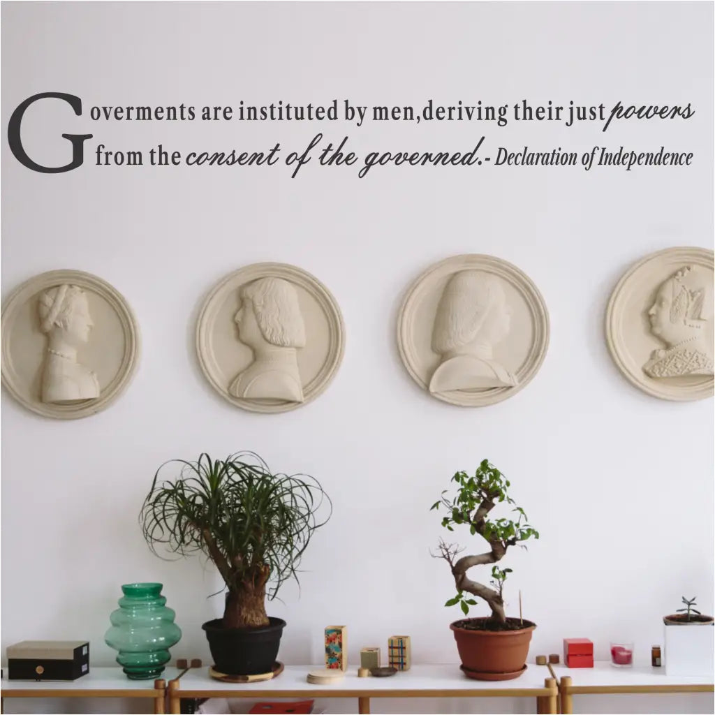 A black wall decal featuring a quote from the Declaration of Independence, "Governments are instituted by men, deriving their just powers from the consent of the governed." The decal is installed in a modern, light-filled room above a shelf decorated with four historical profile reliefs, potted plants, and other items.