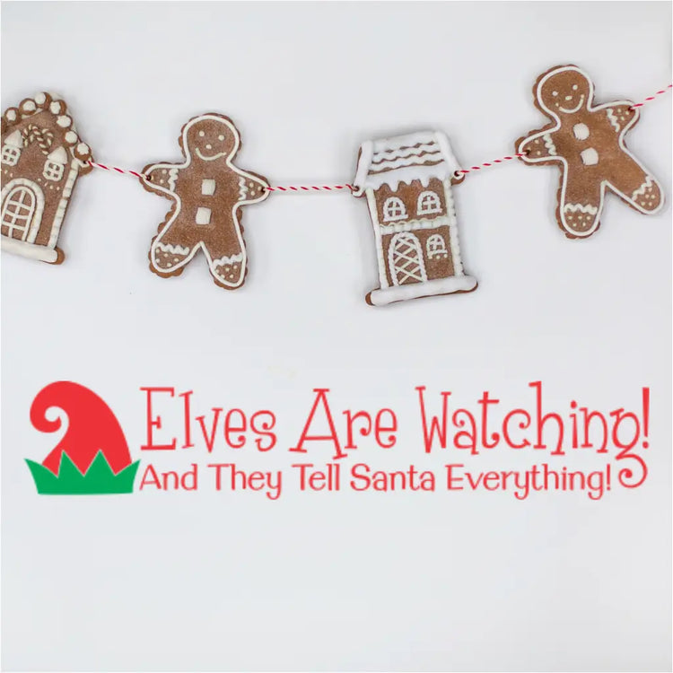 Lifestyle image featuring the red and green Christmas decal 'Elves Are Watching! And They Tell Santa Everything!' underneath a string of gingerbread men and gingerbread house cookies hung on a red and white baker's twine.