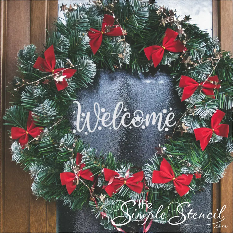 A traditional green evergreen wreath decorated with numerous small red bows and shimmering gold tinsel, hanging on a door with a glass panel. A white "Welcome" decal is prominently displayed in the center of the wreath on the glass.